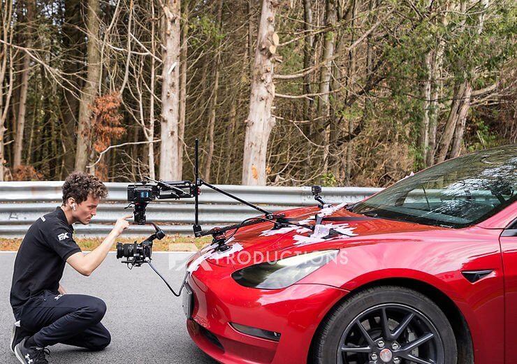 FX Productions Canada A person adjusts a camera rig mounted on the hood of a red car parked on a road near a forested area, showcasing the behind-the-scenes work of an FX Productions Canada video production company. Toronto Video Production Company