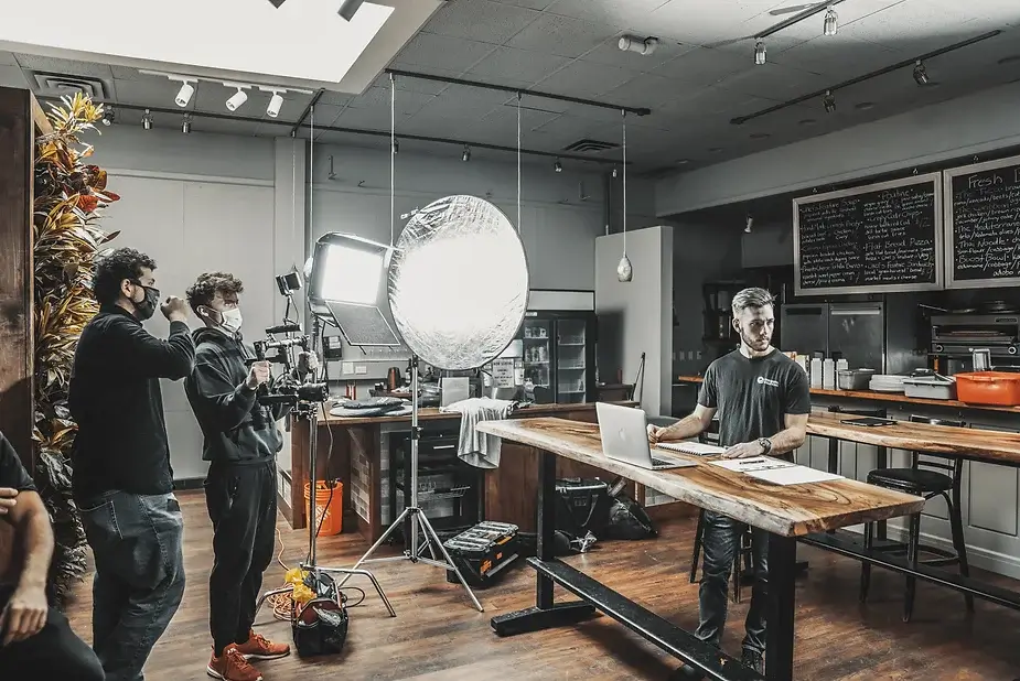 FX Productions Canada A film crew with camera and lighting equipment from Toronto Video Production records a man working on a laptop at a wooden table in a modern kitchen setting. Toronto Video Production Company