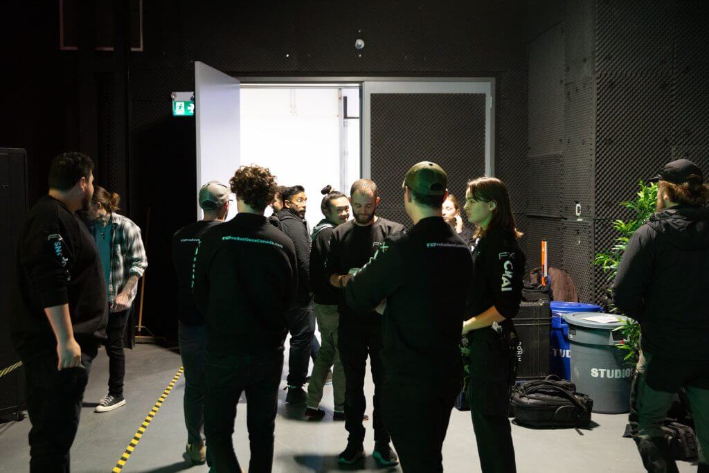 FX Productions Canada A group of people wearing dark clothing stand and talk near an open doorway in an industrial-style room with plants and equipment, reflecting the creative energy of a Toronto Video Production company. Toronto Video Production Company
