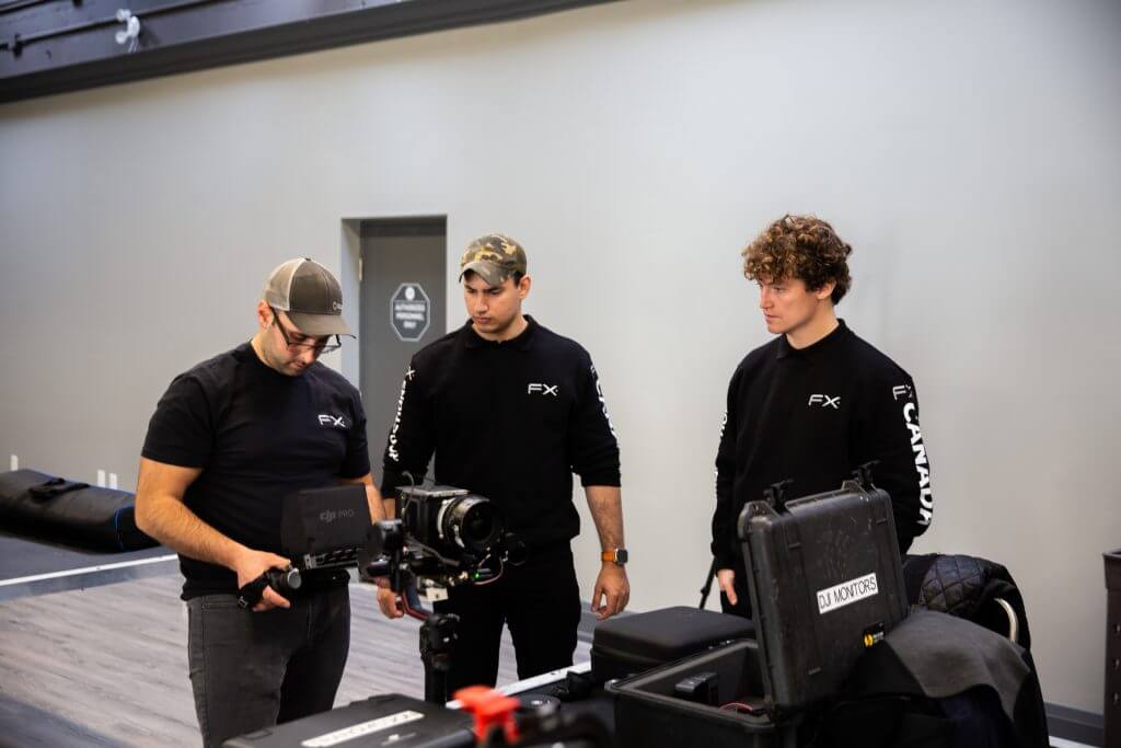FX Productions Canada Three men wearing black FX Productions Canada shirts stand indoors around camera equipment, with one person adjusting a camera while the others observe. Toronto Video Production Company