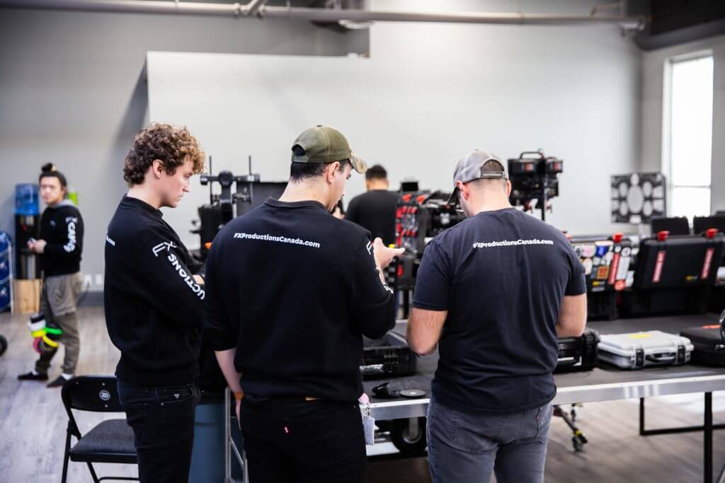 FX Productions Canada Three people in matching shirts stand at a workbench with electronic equipment, focusing on a device; others and gear are visible in the background at FX Productions Canada, a leading video production company. Toronto Video Production Company