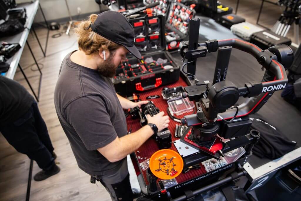 FX Productions Canada A man wearing a cap assembles camera equipment on a table covered with gear, cases, and tools in a well-lit workspace at FX Productions Canada, a leading Toronto Video Production company. Toronto Video Production Company
