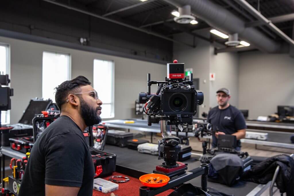 FX Productions Canada A man closely examines a professional video camera setup on a table in a workshop at FX Productions Canada, with another person and equipment visible in the background. Toronto Video Production Company