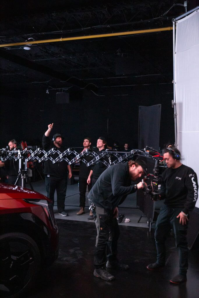 FX Productions Canada Several crew members from FX Productions Canada work on a film set with camera equipment near a red car, some adjusting gear while others observe or point toward the ceiling. Toronto Video Production Company