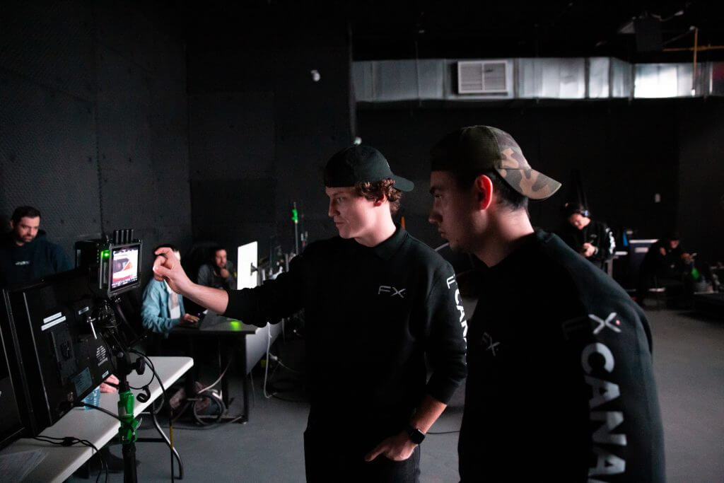 FX Productions Canada Two people wearing black FX CANADA shirts look at a monitor in a dimly lit Toronto Video Production studio, with computers and other team members working in the background. Toronto Video Production Company