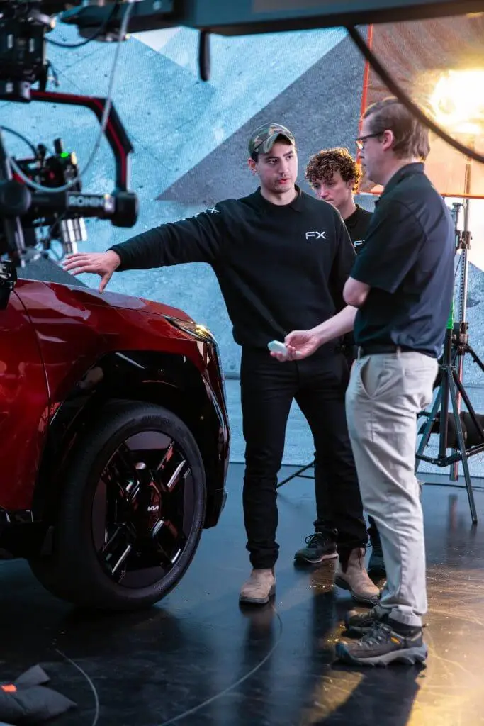 FX Productions Canada Three men stand near a red car on a set with film equipment, discussing something while one points at the vehicle—capturing a behind-the-scenes moment from a Toronto Video Production shoot. Toronto Video Production Company