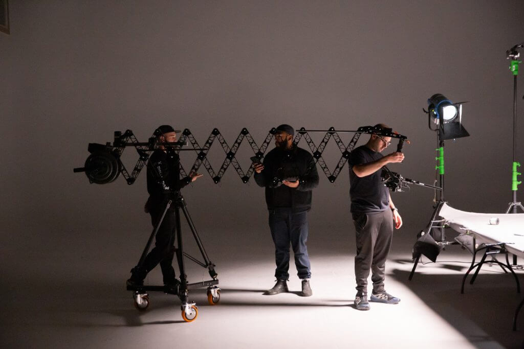 FX Productions Canada Three people from FX Productions Canada set up film equipment and lighting in a studio with a white backdrop; one holds a camera, another adjusts a tripod, and a third stands by a table during this Toronto Video Production session. Toronto Video Production Company
