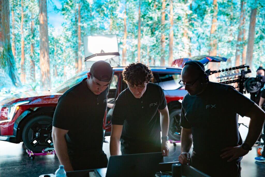 FX Productions Canada Three people in black shirts stand in front of a laptop, collaborating on a film set with a red car and forest background—showcasing the creative work of FX Productions Canada, a leading Toronto Video Production company. Toronto Video Production Company