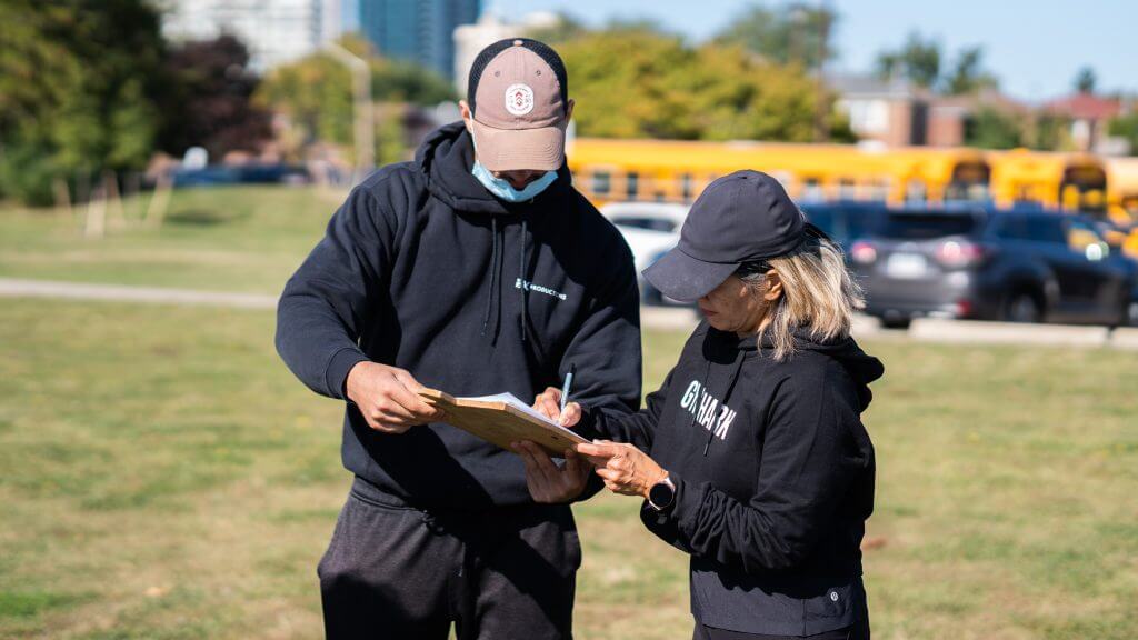 FX Productions Canada Two people wearing black hoodies and caps stand outside on grass, reviewing a clipboard together; a school bus and parked cars are in the background—a candid moment captured by FX Productions Canada, a leading Toronto video production company. Toronto Video Production Company