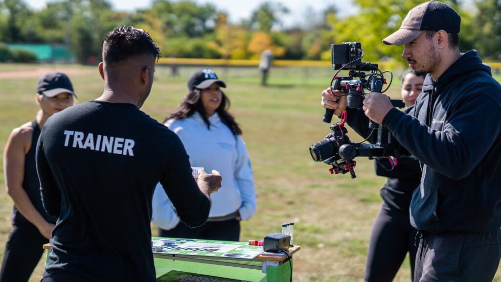 FX Productions Canada A man in a "Trainer" shirt stands at a table outdoors while another films the scene for FX Productions Canada; two women watch in the background. Toronto Video Production Company