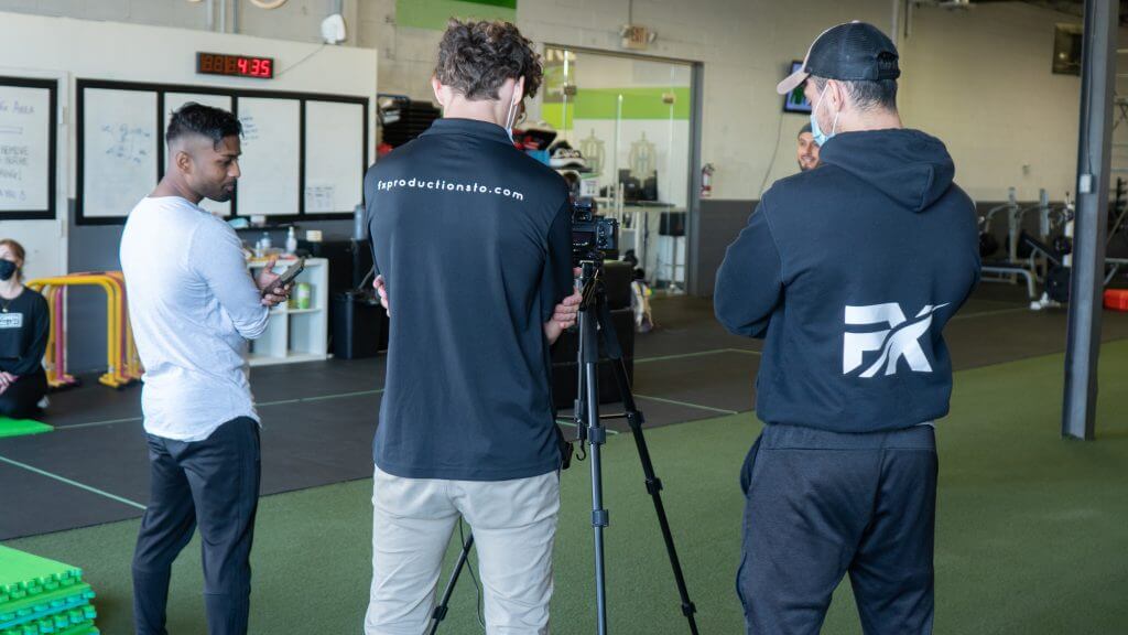 FX Productions Canada Three men stand around a camera tripod in a gym setting, with gym equipment and whiteboards in the background, capturing footage for FX Productions Canada, a leading Toronto Video Production company. Toronto Video Production Company