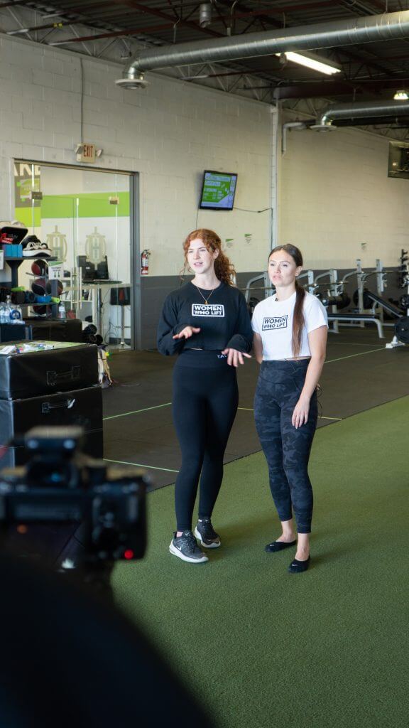 FX Productions Canada Two women stand on artificial turf in a gym, facing the camera. Gym equipment and TV screens fill the background, showcasing a scene that could be captured by a Toronto Video Production company. Toronto Video Production Company