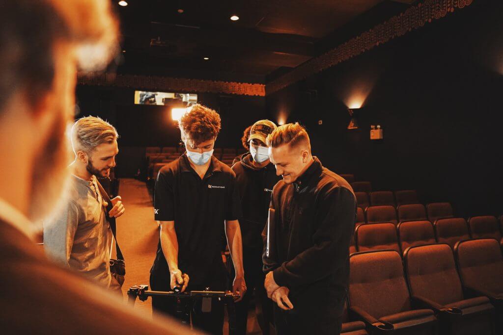 FX Productions Canada Four people stand in a dimly lit auditorium, two wearing masks and handling camera equipment for FX Productions Canada, while the others observe. Empty seats fill the background. Toronto Video Production Company