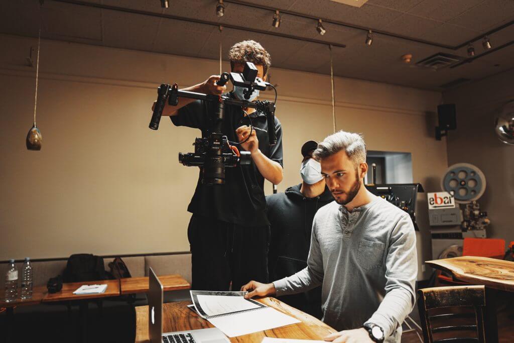 FX Productions Canada A man works on a laptop at a desk while two crew members from FX Productions Canada stand behind him, operating a camera on a stabilizer in an indoor Toronto video production setting. Toronto Video Production Company