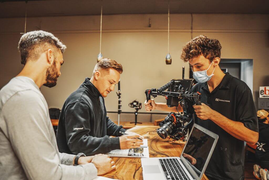 FX Productions Canada Three men work at a table; two review documents while the third, wearing a mask, adjusts a camera rig beside a laptop at FX Productions Canada, a leading Toronto video production company. Toronto Video Production Company