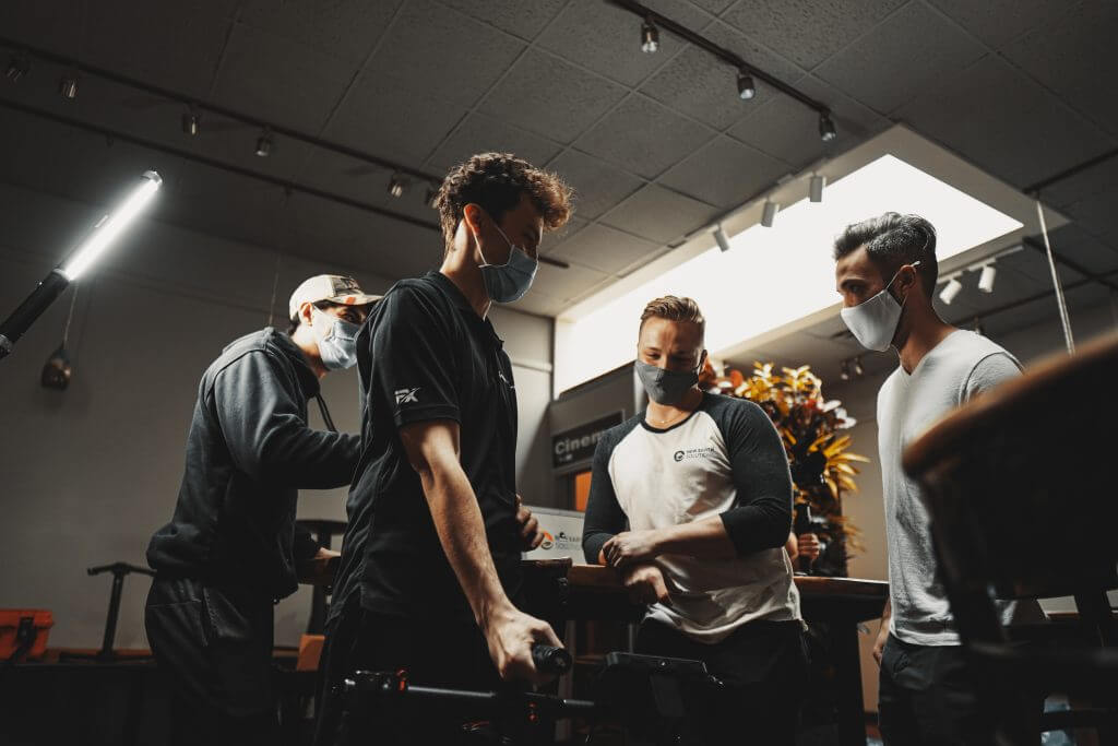 FX Productions Canada Four men wearing face masks stand together indoors, engaged in conversation around a table under bright ceiling lights at FX Productions Canada, a leading Toronto Video Production company. Toronto Video Production Company