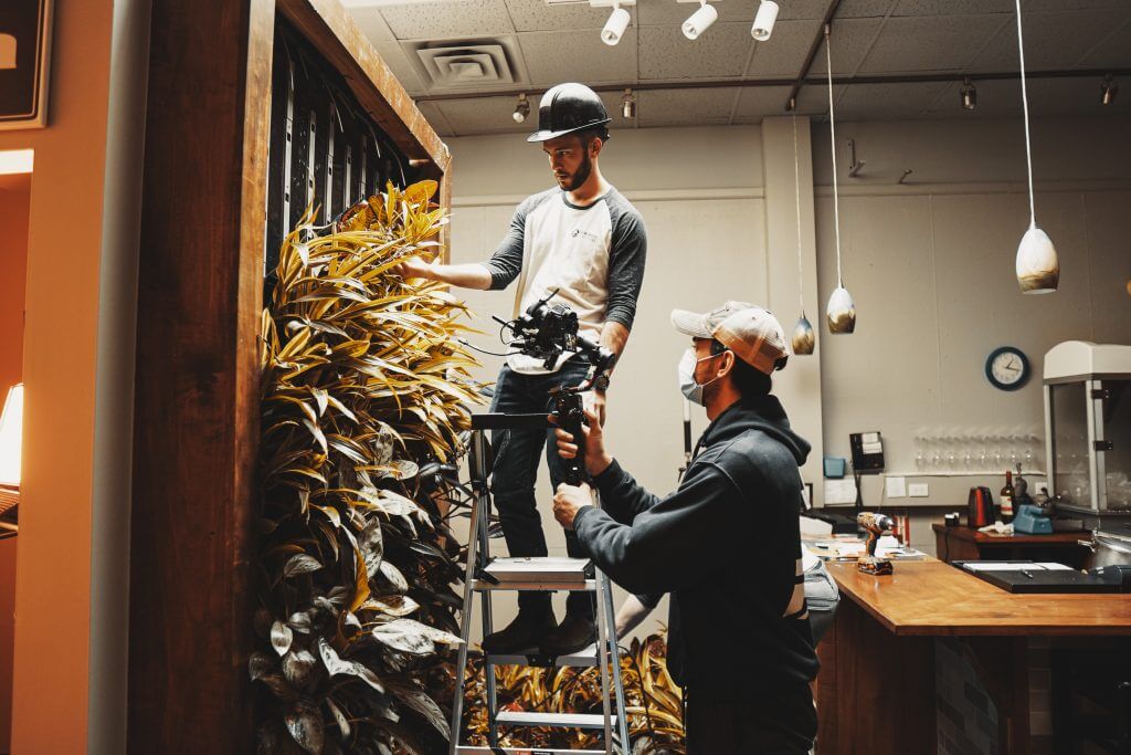 FX Productions Canada Two men work indoors; one stands on a ladder adjusting plants on a wall while the other, wearing a mask, films him with a camera for FX Productions Canada. Toronto Video Production Company