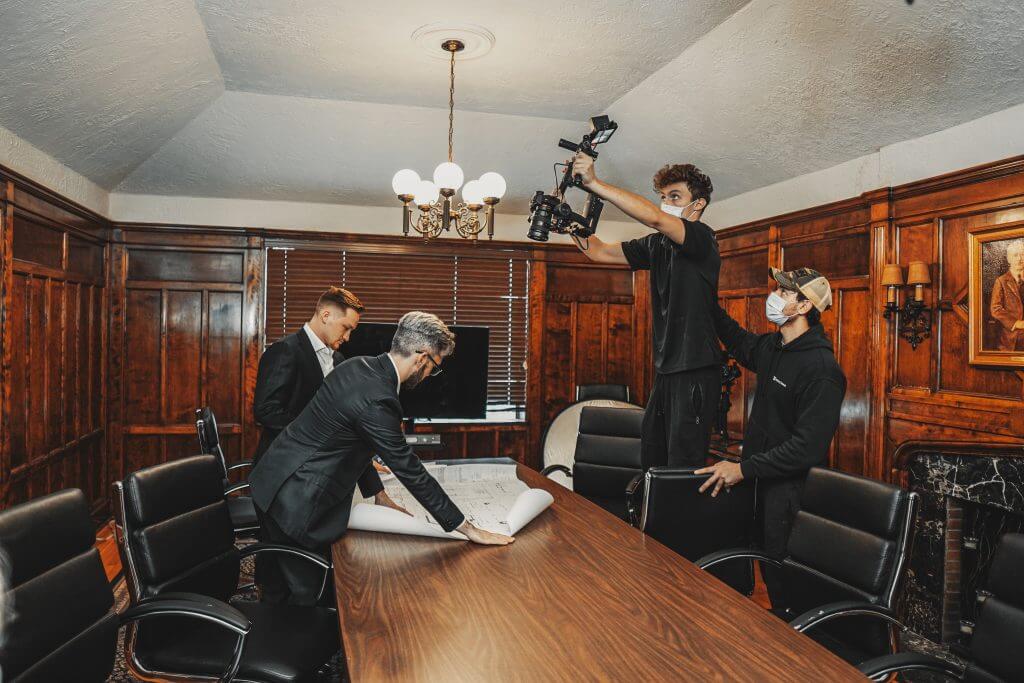 FX Productions Canada Two men in suits review documents at a conference table while two crew members from FX Productions Canada film them, one holding a camera above his head in the wood-paneled meeting room. Toronto Video Production Company