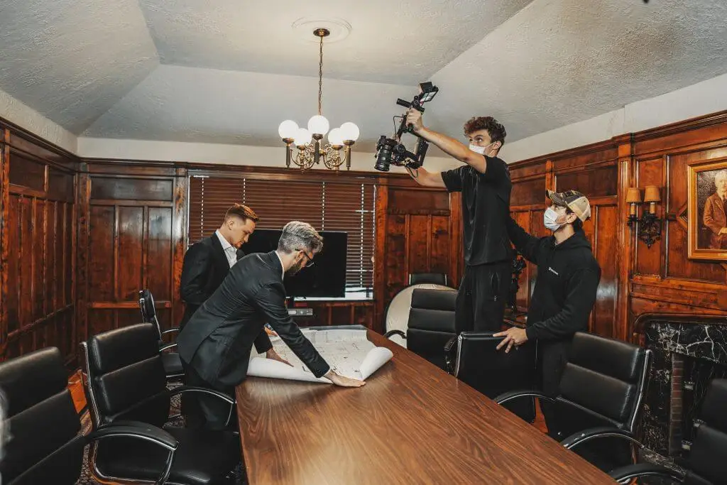 FX Productions Canada Two men in suits review documents at a conference table while two crew members from FX Productions Canada film them, one holding a camera above his head in the wood-paneled meeting room. Toronto Video Production Company