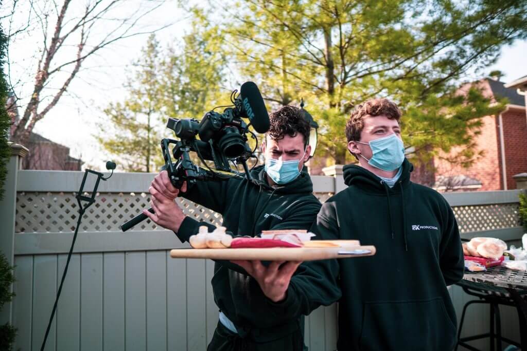 FX Productions Canada Two men wearing masks stand outside; one holds a camera rig while the other presents a tray of food. Trees and a fence are visible in the background, highlighting an on-location Toronto Video Production shoot. Toronto Video Production Company