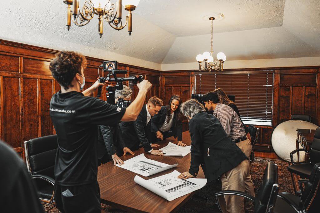 FX Productions Canada A group of people in business attire reviews architectural plans on a table while a person from a Toronto Video Production company films them in a wood-paneled conference room. Toronto Video Production Company