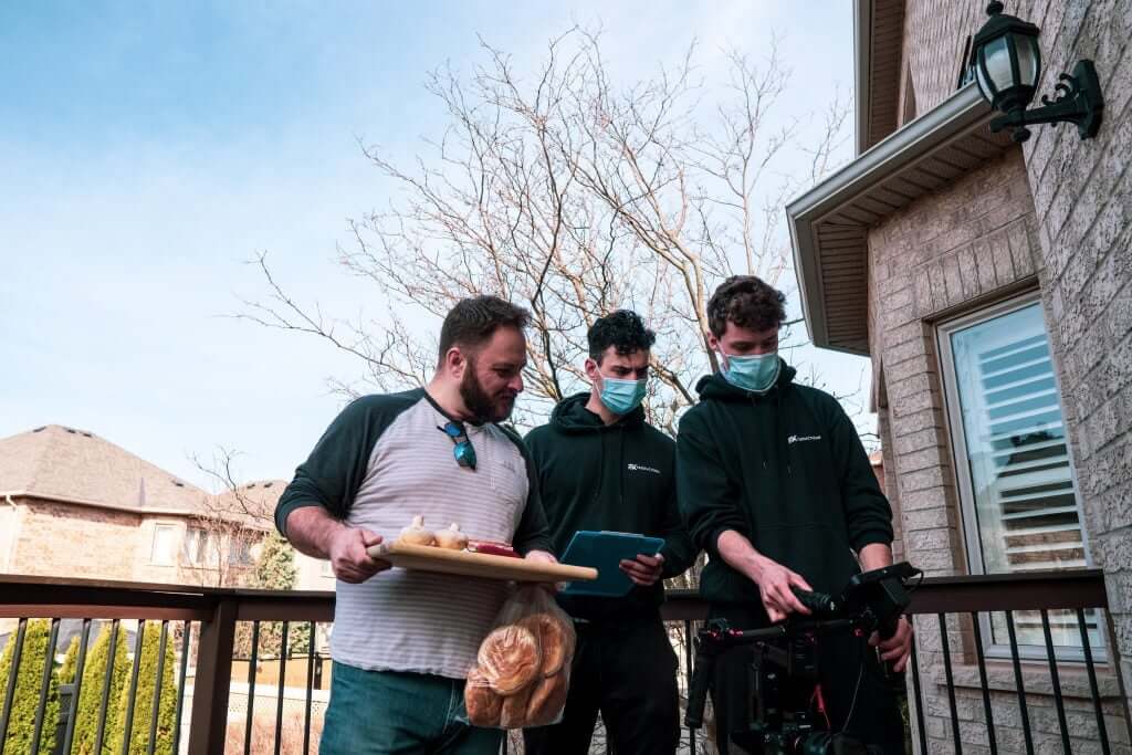 FX Productions Canada Three men stand on a deck; one holds a tray with bread and snacks while the other two, wearing face masks, look at a Toronto Video Production camera setup. Toronto Video Production Company