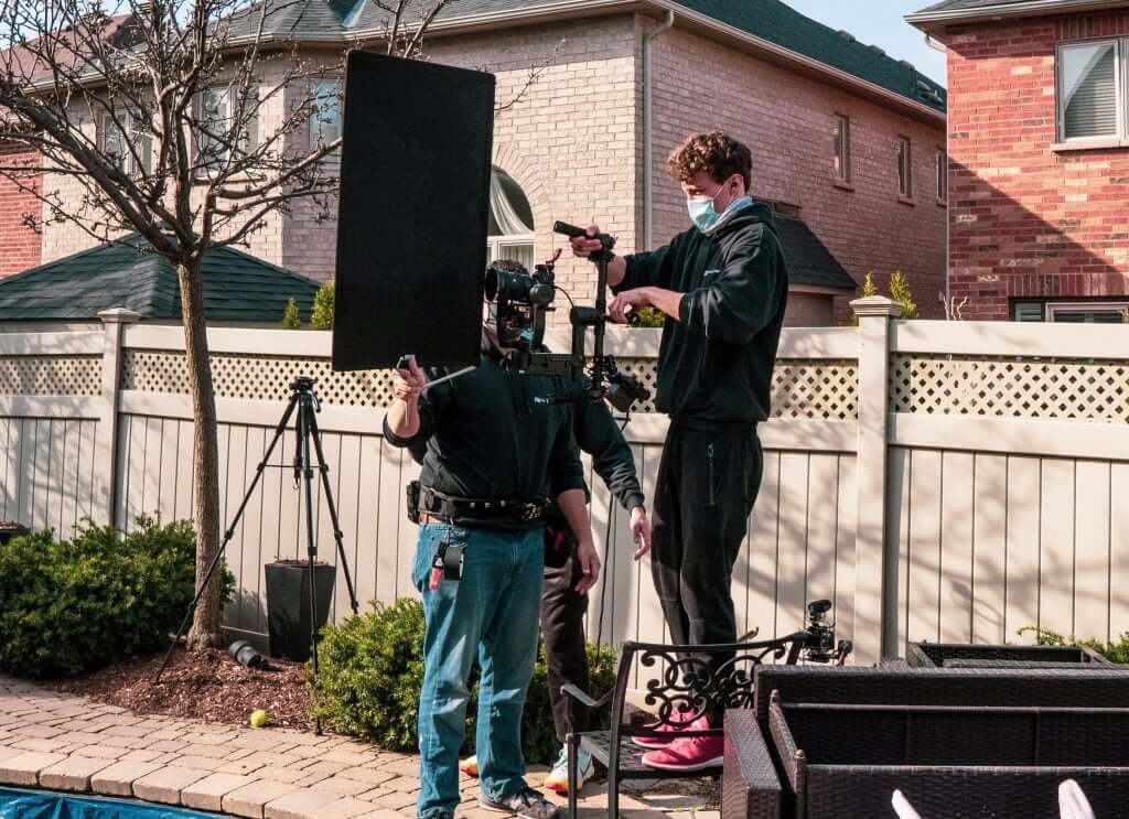 FX Productions Canada Two people from a Toronto Video Production company set up film equipment in a backyard, one holding a camera rig and the other a large reflector. Both are wearing masks. Toronto Video Production Company