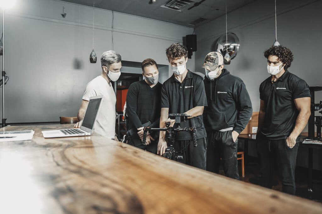 FX Productions Canada Five people wearing masks stand indoors around a camera setup on a tripod, with a laptop on a wooden table in the foreground—an inside look at Toronto Video Production by FX Productions Canada. Toronto Video Production Company