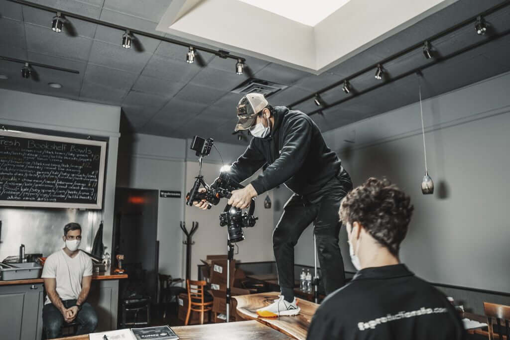 FX Productions Canada A person wearing a mask and cap stands on a table with a camera rig, filming another masked individual while a third person sits in the background—capturing the creative process at FX Productions Canada, a leading Toronto Video Production company. Toronto Video Production Company