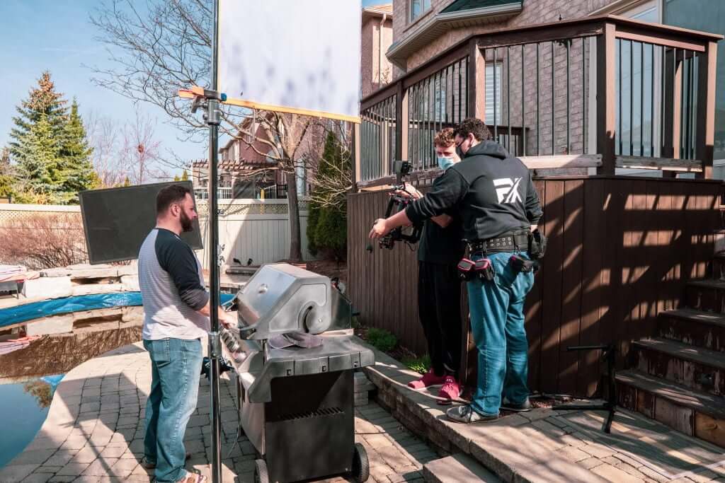 FX Productions Canada Three people from a Toronto Video Production team film a man cooking at a grill in a backyard. The cameraman and sound operator wear equipment, and one person is wearing a face mask. Toronto Video Production Company