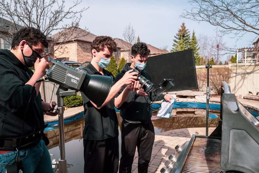 FX Productions Canada Three people wearing masks adjust camera and lighting equipment next to an outdoor grill in a backyard setting, capturing the behind-the-scenes work of a Toronto Video Production team. Toronto Video Production Company