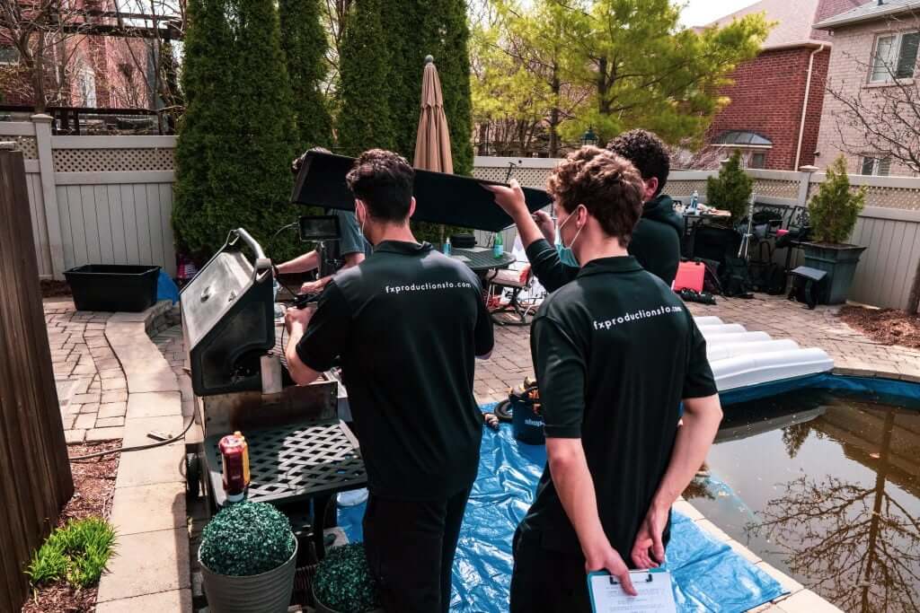 FX Productions Canada Three people in matching shirts with “tspprofessionalclean.com” on the back work together on a grill in a backyard near a pond, as tools and a notepad sit nearby—captured by Toronto Video Production for authentic team moments. Toronto Video Production Company