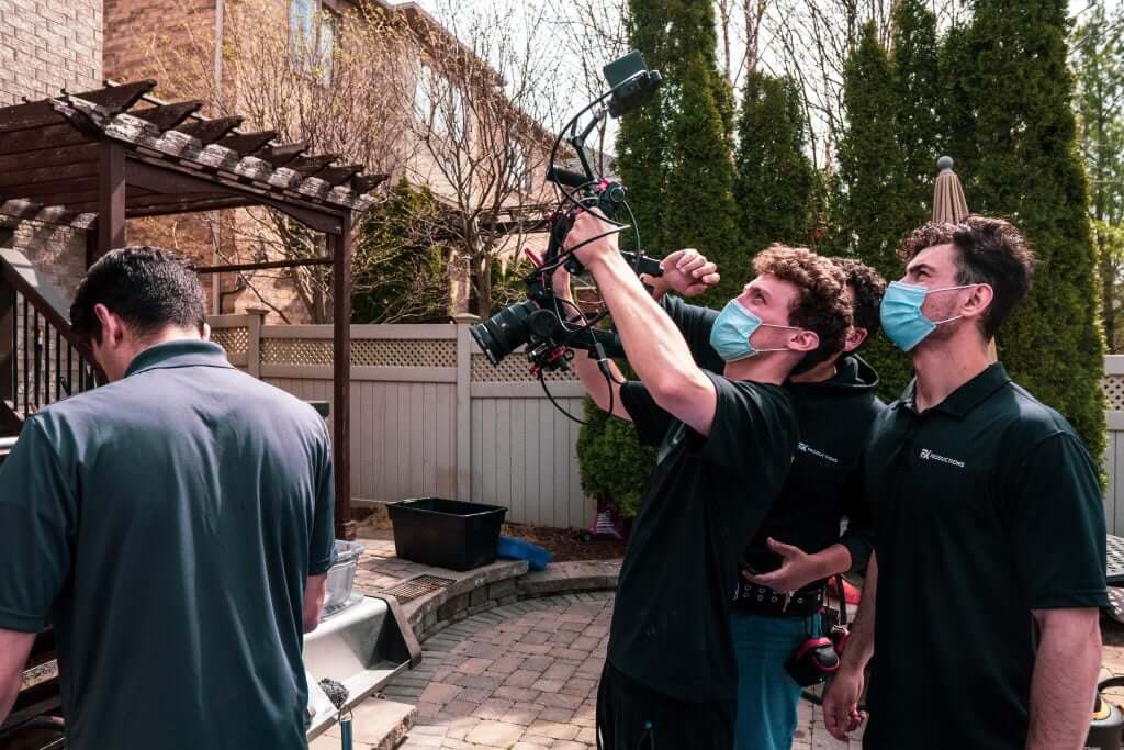 FX Productions Canada Three people in masks and black shirts are filming outdoors for FX Productions Canada; one operates a camera rig while the others observe, all standing in a backyard with trees and a wooden pergola—a glimpse into Toronto Video Production. Toronto Video Production Company