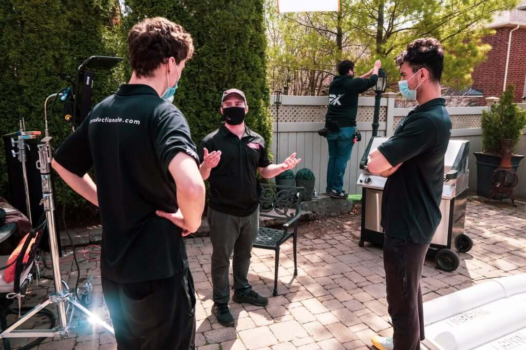 FX Productions Canada Three people in black shirts and masks talk outdoors near film equipment, while a fourth person adjusts something on a fence in the background during a Toronto Video Production shoot. Toronto Video Production Company