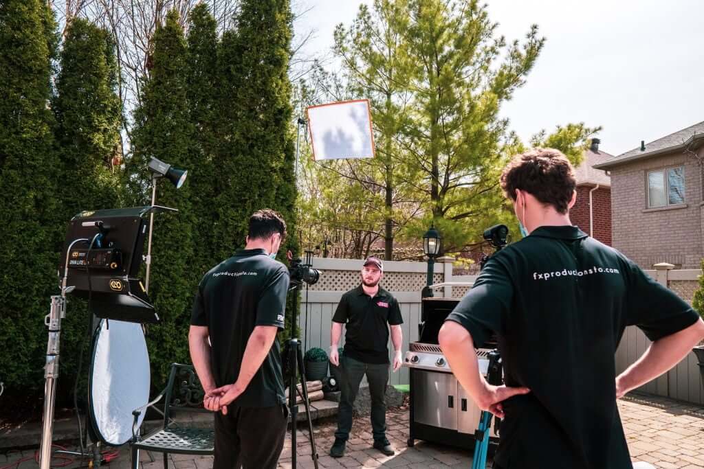 FX Productions Canada Three crew members from a Toronto Video Production company film a person standing next to a grill in a backyard, using professional video and lighting equipment on a sunny day. Toronto Video Production Company