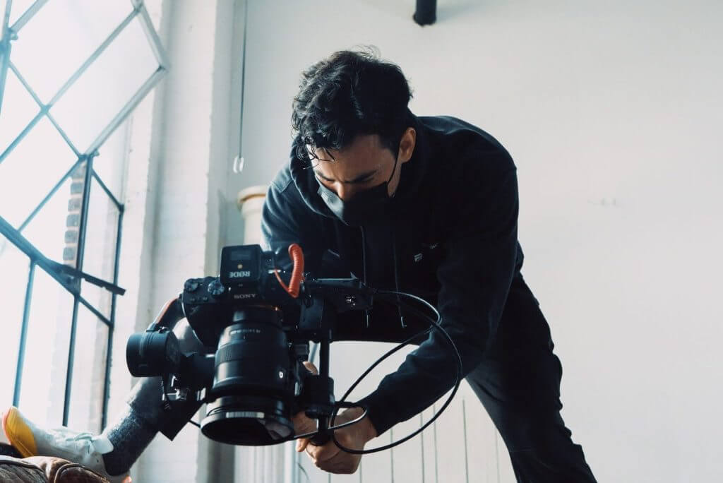 FX Productions Canada A person wearing a black mask and hoodie is operating a professional video camera indoors near large windows, capturing dynamic shots for FX Productions Canada. Toronto Video Production Company