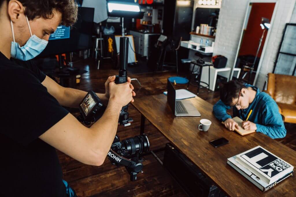 FX Productions Canada A cameraman from FX Productions Canada films a person writing in a notebook at a wooden table in a studio setting, with film equipment and books visible—a glimpse into Toronto Video Production behind the scenes. Toronto Video Production Company