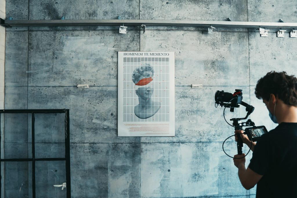 FX Productions Canada A person films a poster of a classical statue head with an orange band over its eyes, mounted on a concrete wall—showcasing the creative eye of Toronto Video Production experts. Toronto Video Production Company