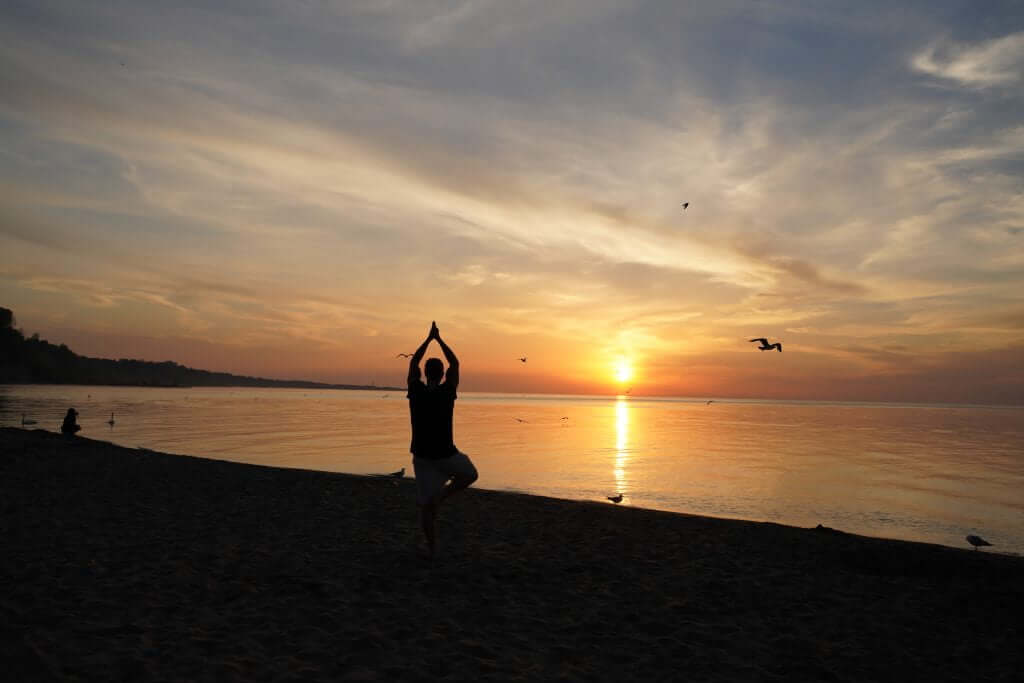 FX Productions Canada Person practicing yoga in a tree pose on a beach at sunset, with calm water, birds flying, and an orange sky in the background—captured beautifully by FX Productions Canada, your go-to Toronto video production company. Toronto Video Production Company
