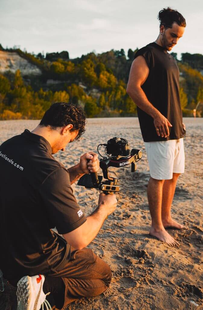 FX Productions Canada A man kneels on sand operating a camera stabilizer while another stands barefoot nearby, both outdoors with trees and hills behind them—a dynamic moment captured by FX Productions Canada, a leading Toronto Video Production company. Toronto Video Production Company
