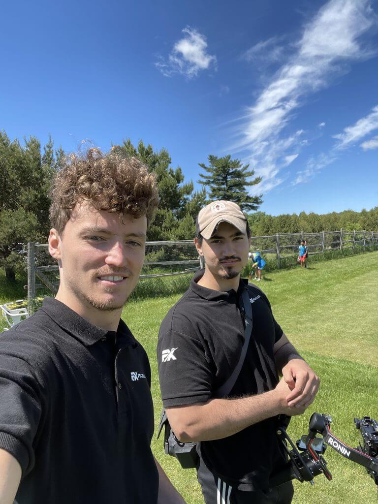 FX Productions Canada Two men in black polo shirts stand on grass with camera equipment, trees, a fence, and blue sky with clouds behind them—capturing the essence of a Toronto Video Production company in action. Toronto Video Production Company