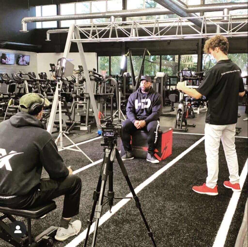 FX Productions Canada A man is being interviewed in a gym, sitting on a red box while two crew members from FX Productions Canada film and record him with professional video and audio equipment. Toronto Video Production Company