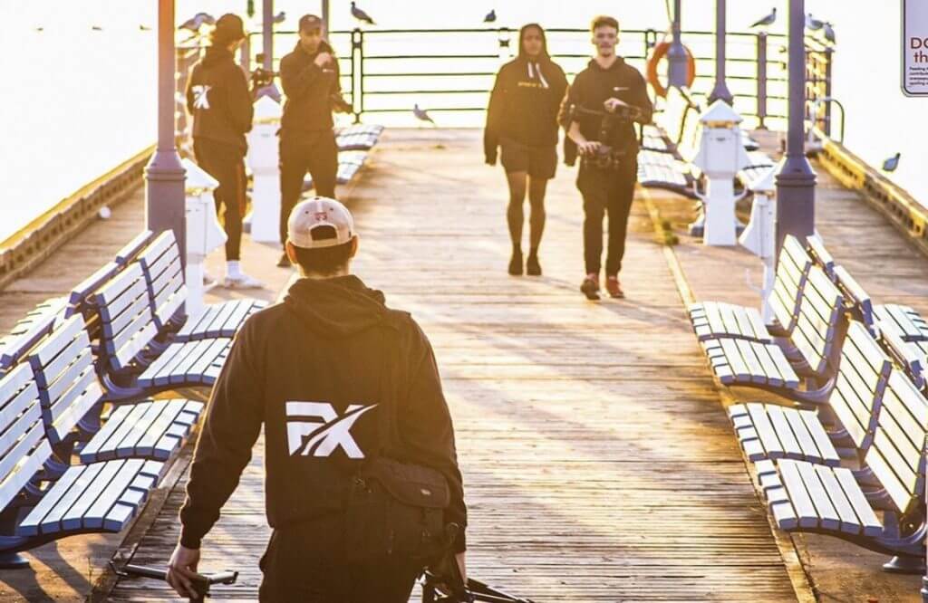 FX Productions Canada A group of people, some holding camera equipment from FX Productions Canada, walk along a wooden pier lined with blue benches and railings on a sunny day. Toronto Video Production Company