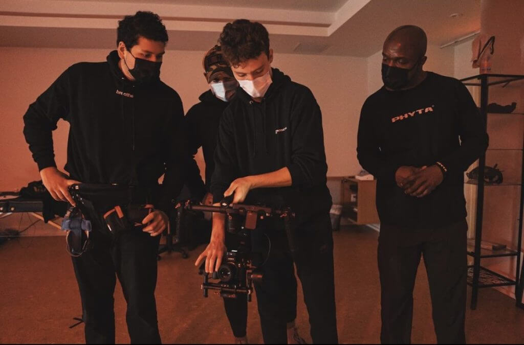 FX Productions Canada Four people wearing black clothing and face masks stand indoors, closely examining and adjusting professional camera equipment for a Toronto Video Production project. Toronto Video Production Company