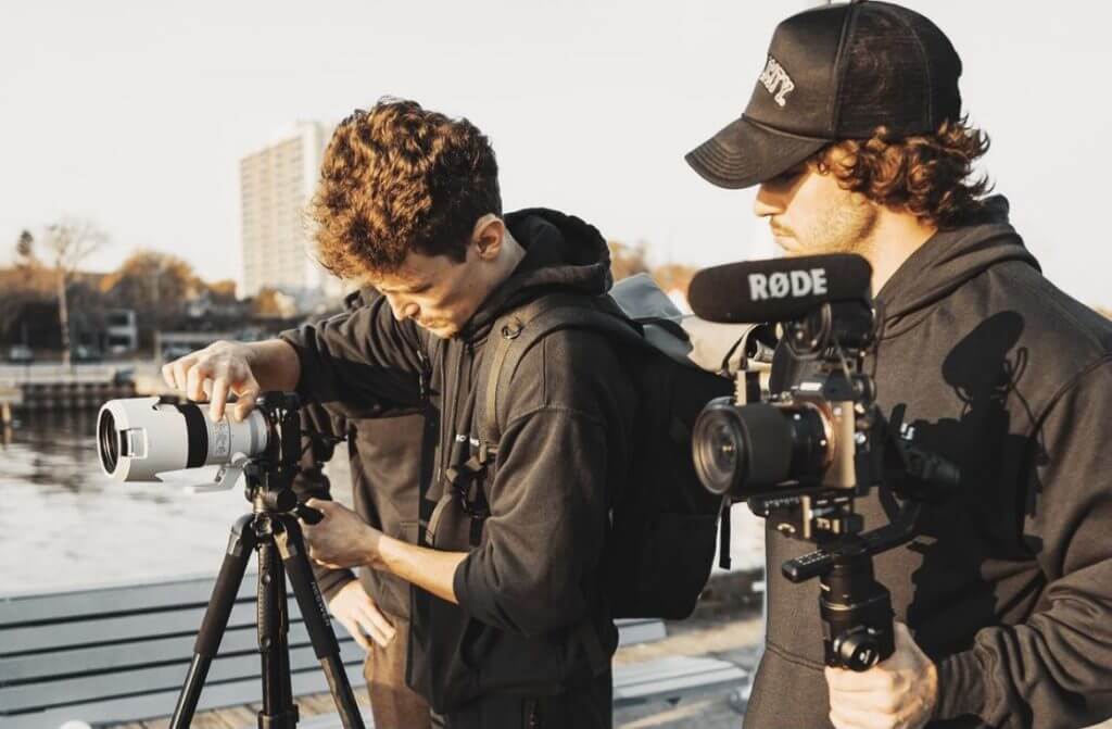 FX Productions Canada Two people in black hoodies operate cameras by the water; one adjusts a camera on a tripod while the other holds a camera with a RODE microphone, showcasing behind-the-scenes action from Toronto Video Production experts. Toronto Video Production Company