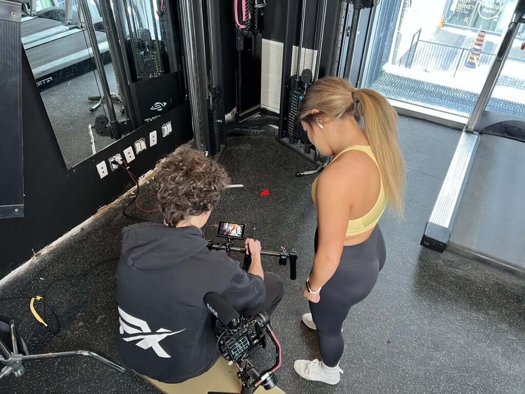 FX Productions Canada A man operates a camera on the gym floor while a woman in athletic wear stands nearby, looking down at the camera screen, during a Toronto Video Production shoot. Toronto Video Production Company
