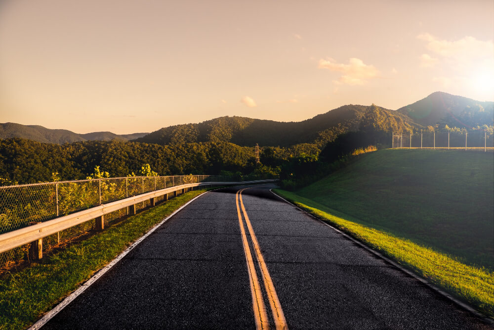 FX Productions Canada A paved road with double yellow lines curves through green hills and trees, with mountains and a clear sky in the background at sunset—an inspiring scene perfect for an FX Productions Canada shoot. Toronto Video Production Company