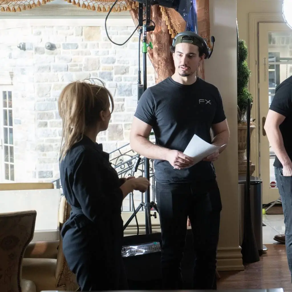FX Productions Canada Two people stand indoors having a conversation; one holds papers and wears a black FX Productions Canada t-shirt and cap, while film equipment from a Toronto Video Production company is visible in the background. Toronto Video Production Company