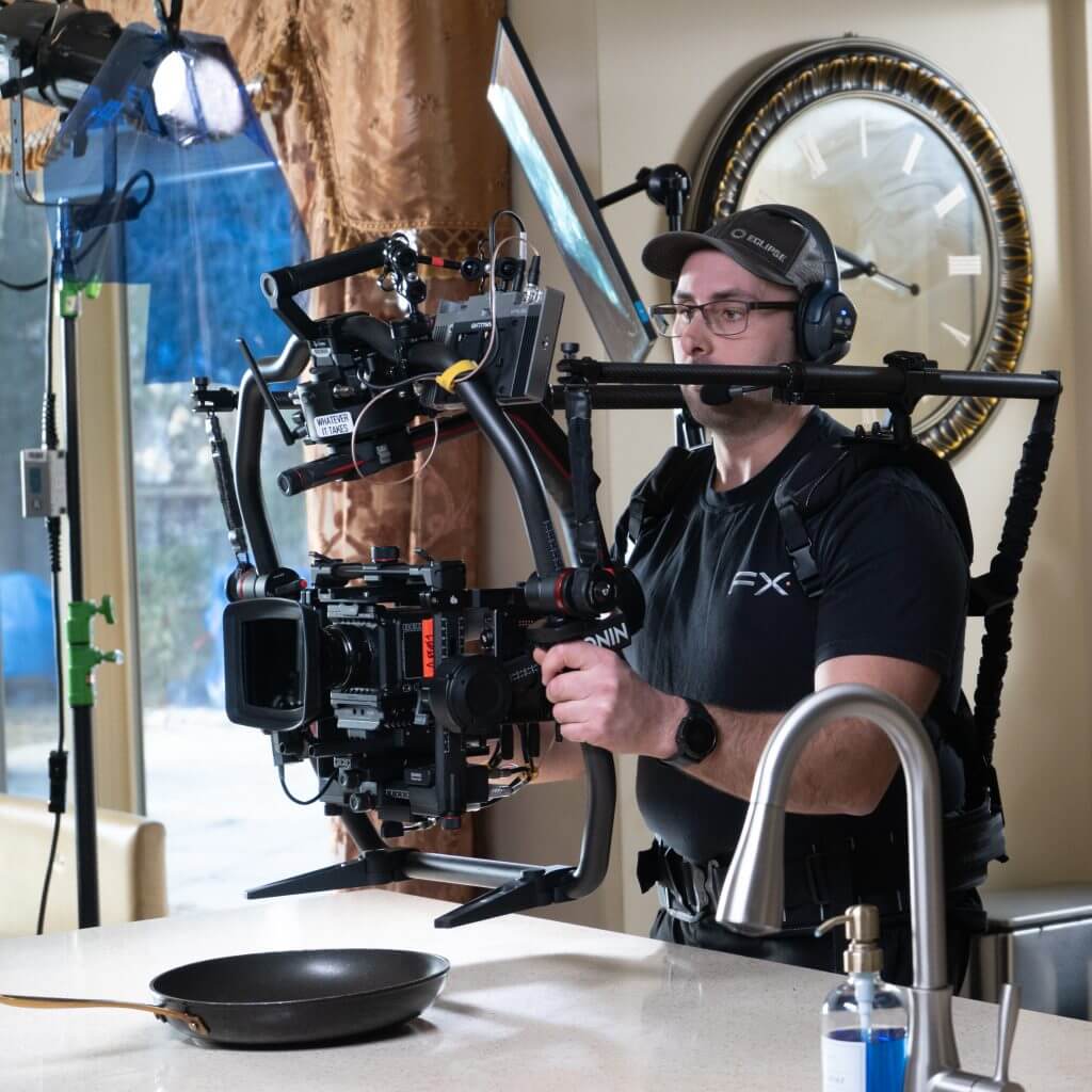 FX Productions Canada A camera operator from FX Productions Canada, wearing a headset and cap, films a kitchen scene with a professional camera rig, standing behind a counter with a frying pan and soap dispenser. Toronto Video Production Company