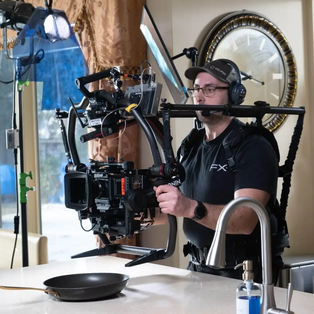 FX Productions Canada A camera operator from FX Productions Canada, wearing a headset and cap, films a kitchen scene with a professional camera rig, standing behind a counter with a frying pan and soap dispenser. Toronto Video Production Company